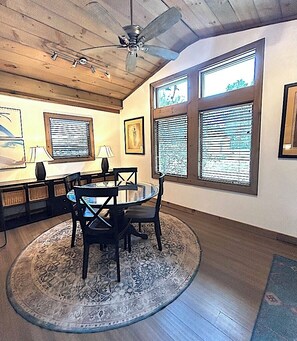 A dining area with a round table, chairs, a ceiling fan, and large windows with blinds. - Welcome to Little Bit O' the Bay (Saint Leonard)