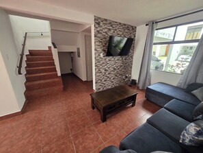 A living room with a staircase, a flat-screen TV mounted on a textured wall, a dark sofa, and a coffee table. - Just 15 minutes from Antigua Guatemala, within the La Joya Condominium. (Santa Lucía Milpas Altas)