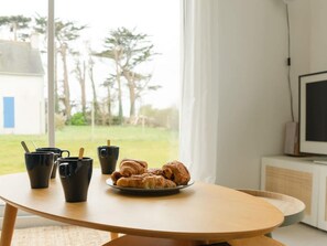 A table with black cups and a plate of croissants, a TV, and a window with a view of trees and a building. - Charming Coastal Home in Landunvez (Landunvez)
