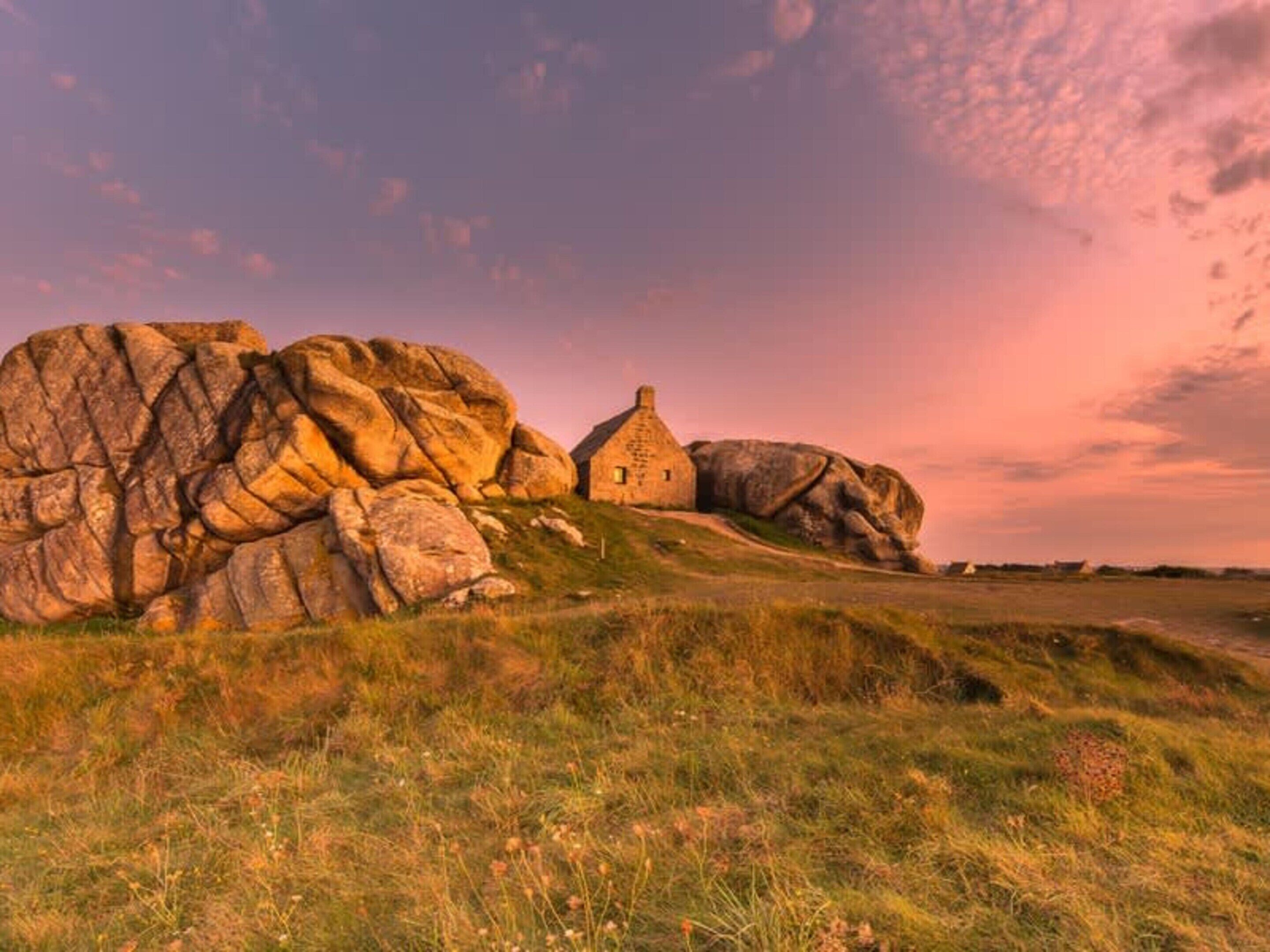 A stone house with a thatched roof, surrounded by large boulders and grassy terrain.
