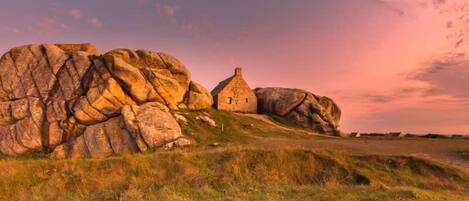 A stone house with a thatched roof, surrounded by large boulders and grassy terrain.
