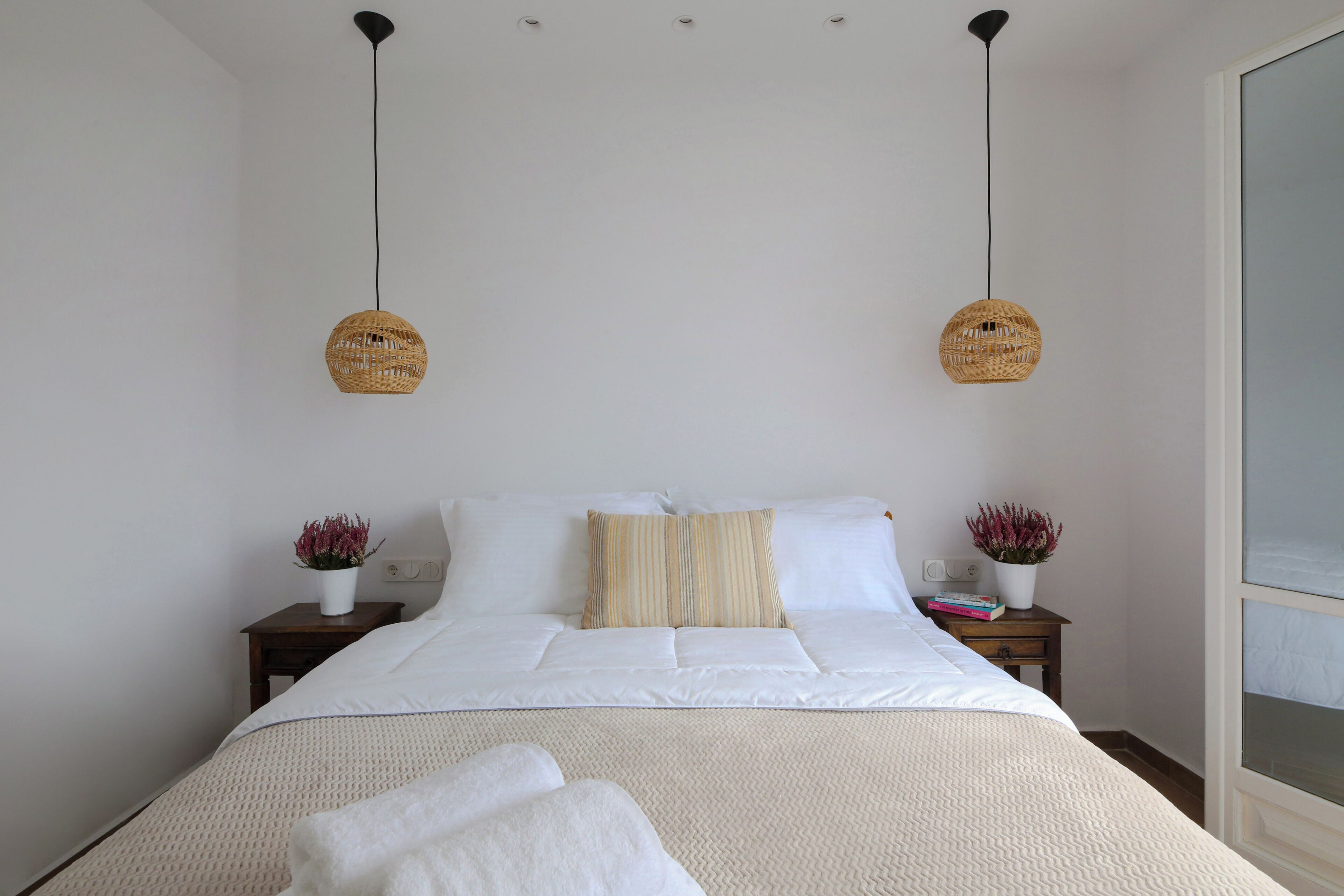 Una cama bien tendida con sábanas blancas, dos mesitas de noche con jarrones decorativos y un ventanal grande a la derecha.