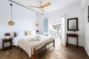 Ein Schlafzimmer mit Bett, Nachttischen, Deckenventilator, Fenster mit Blick und einem kleinen Tisch mit Pflanze.