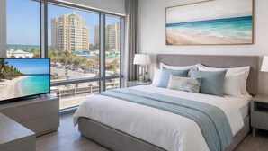 A hotel room with a large bed, a TV on a stand, a painting of a beach, and a view of tall buildings and a beach through the window. - Comfortable King Suite (Clearwater)