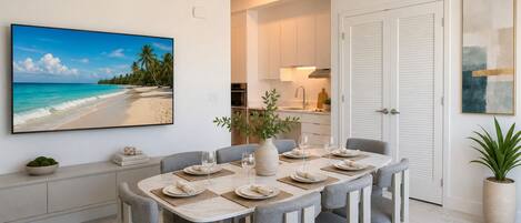 A dining area with a table set for four, a kitchen in the background, and a beach scene on the wall.