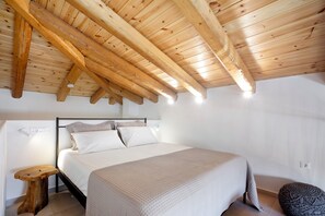 A bedroom with a wooden ceiling, a bed with white bedding, a small wooden stool, and a round ottoman. - Olive Dryopis Apartments (Kithnos)