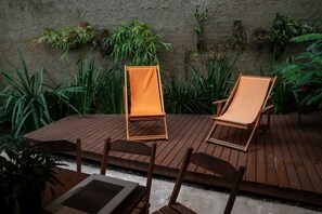 Eine Holzterrasse mit zwei Liegestühlen und einem Tisch, umgeben von üppiger Bepflanzung und einer strukturierten Wand.
