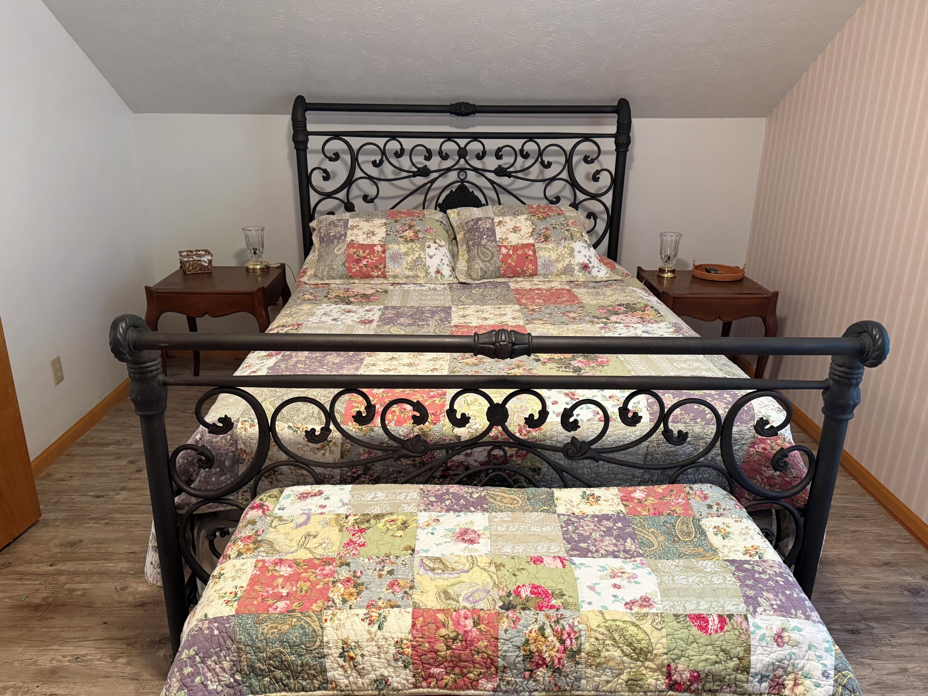 A double bed with a floral quilt, a metal headboard, two bedside tables, and a wooden floor.