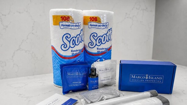 Un conjunto de artículos de aseo que incluye dos rollos de papel higiénico Scott, una caja azul con la marca Marco Island, una tarjeta de bienvenida y un juego de cápsulas para lavadora.
