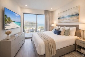 A modern hotel room with a large bed, a TV, a balcony with a view of the beach and hotel, and a painting on the wall. - Relaxed Living Suite (Clearwater)