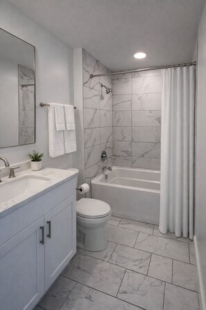 Un baño con ducha revestida de mármol, bañera, inodoro, lavamanos con espejo y una planta en maceta.