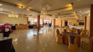 A spacious banquet hall with round tables and chairs, a buffet area, and a large window providing natural light.
