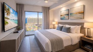 Habitación de hotel moderna con una cama grande, televisión, ventilador de techo y vistas a la playa y edificios.