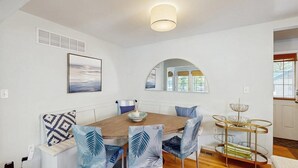 Ein Essbereich mit Holztisch, Stühlen mit blattgemusterten Bezügen, einer weißen Couch mit gemusterten Kissen, einem Wandbild, einem Spiegel und einem Metallständer mit einer Schale.