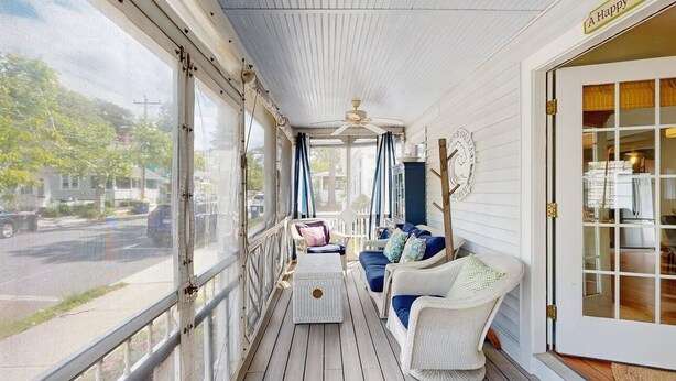 Ein überdachter Balkon mit weißer Decke, einem Deckenventilator, einer Couch mit Kissen und einem Korbsessel.