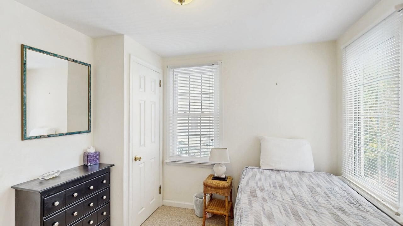 Quarto com cama, cômoda, espelho, janela com persianas e abajur.