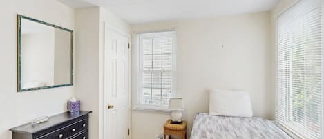 Quarto com cama, cômoda, espelho, janela com persianas e abajur.