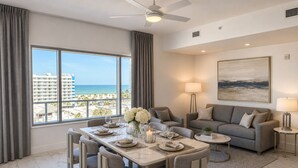 A modern living room with a dining area, large windows with a view of the beach, a ceiling fan, and a painting on the wall. - Coastal Luxe Suite Upper Floor (Clearwater)