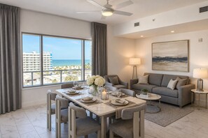 Una sala de estar moderna con comedor, ventanas grandes que ofrecen vistas a la playa, un ventilador de techo y un cuadro en la pared.