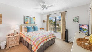 Un dormitorio con cama, mesita de noche, ventilador de techo, ventana con cortinas y televisor.