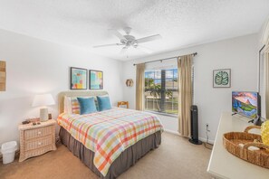 Ein Schlafzimmer mit Bett, Nachttisch, Deckenventilator, Fenster mit Vorhängen und Fernseher.