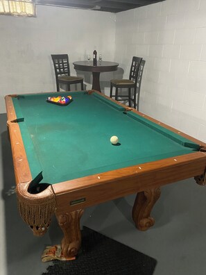Ein Billardtisch mit einer weißen Kugel, einem Satz bunter Kugeln und einem Billardqueue. Zwei Stühle und ein kleiner Tisch mit einer Flasche und Gläsern.