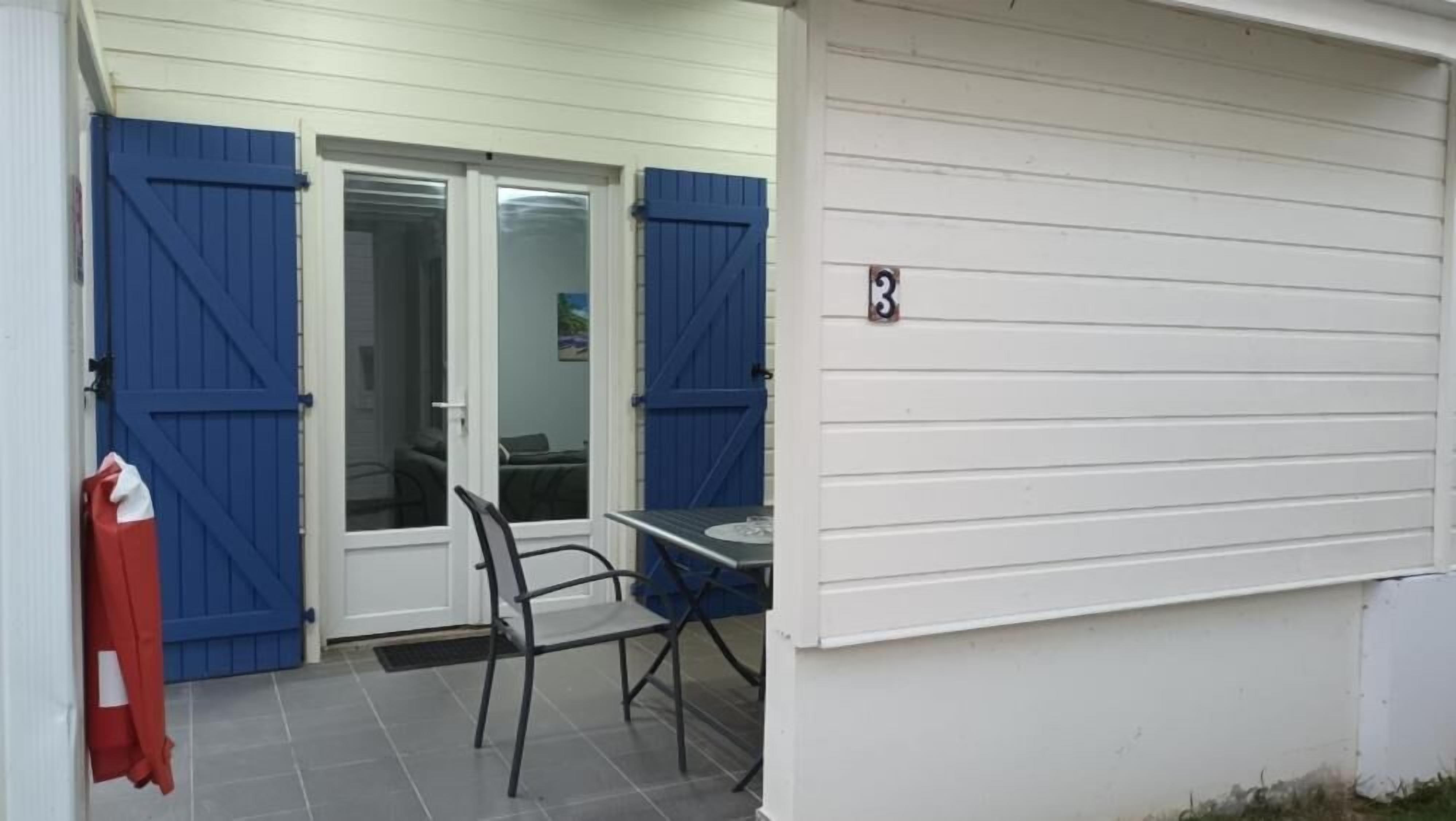 A building with a white exterior, a blue door, a table with a chair, and a red object on the left.