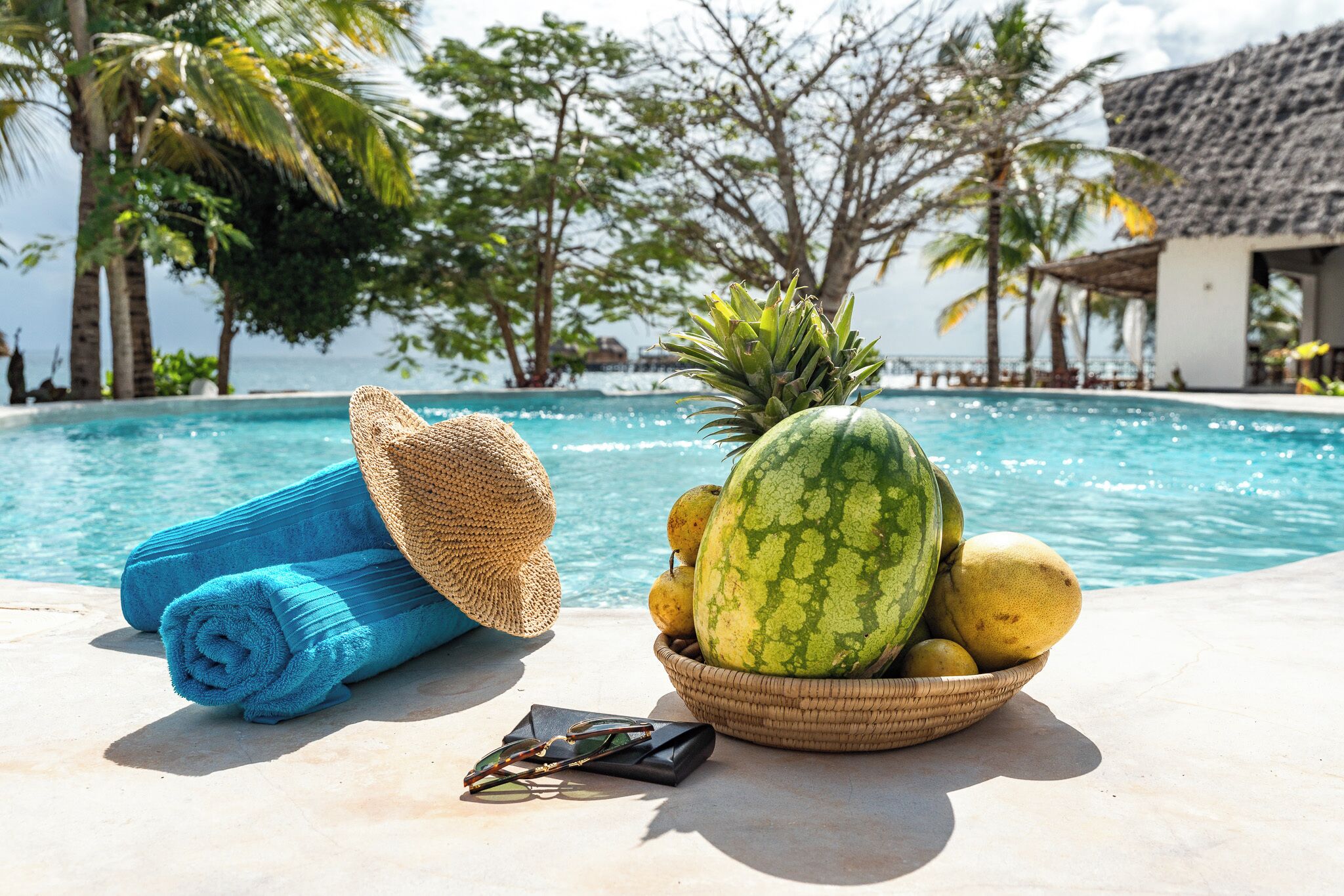 Escena junto a la piscina con un cuenco de fruta, un sombrero de paja y toallas enrolladas.