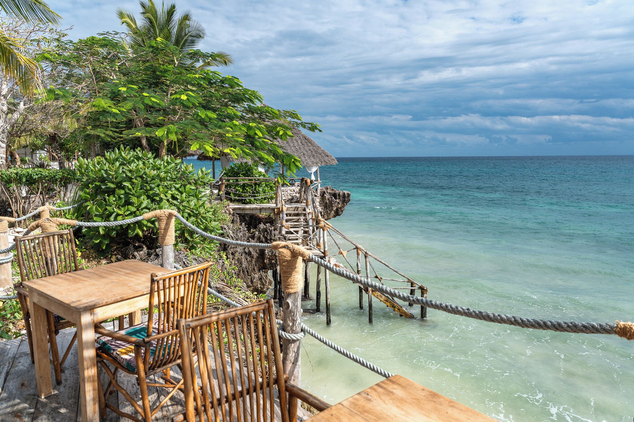 A wooden deck with chairs overlooking turquoise ocean waters, a thatched-roof structure, and lush greenery.