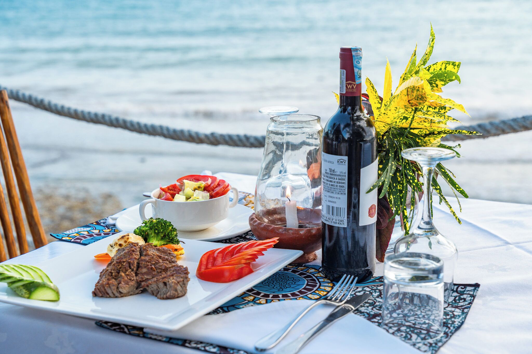 A table with a meal, a bottle of wine, and a candle, set against a beach backdrop.