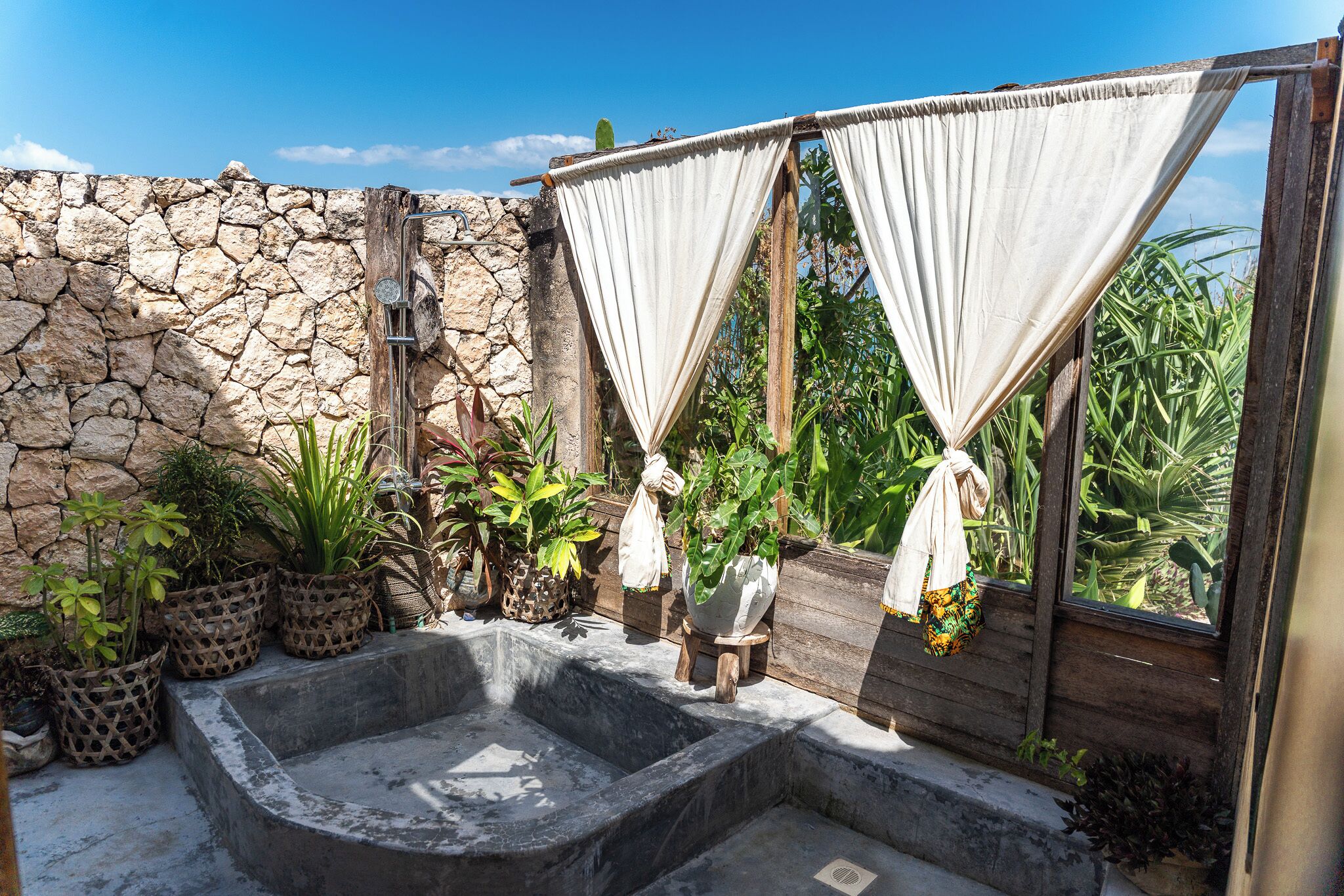 Un espacio al aire libre con paredes de piedra, un jacuzzi, plantas en macetas y una estructura de madera con cortinas.