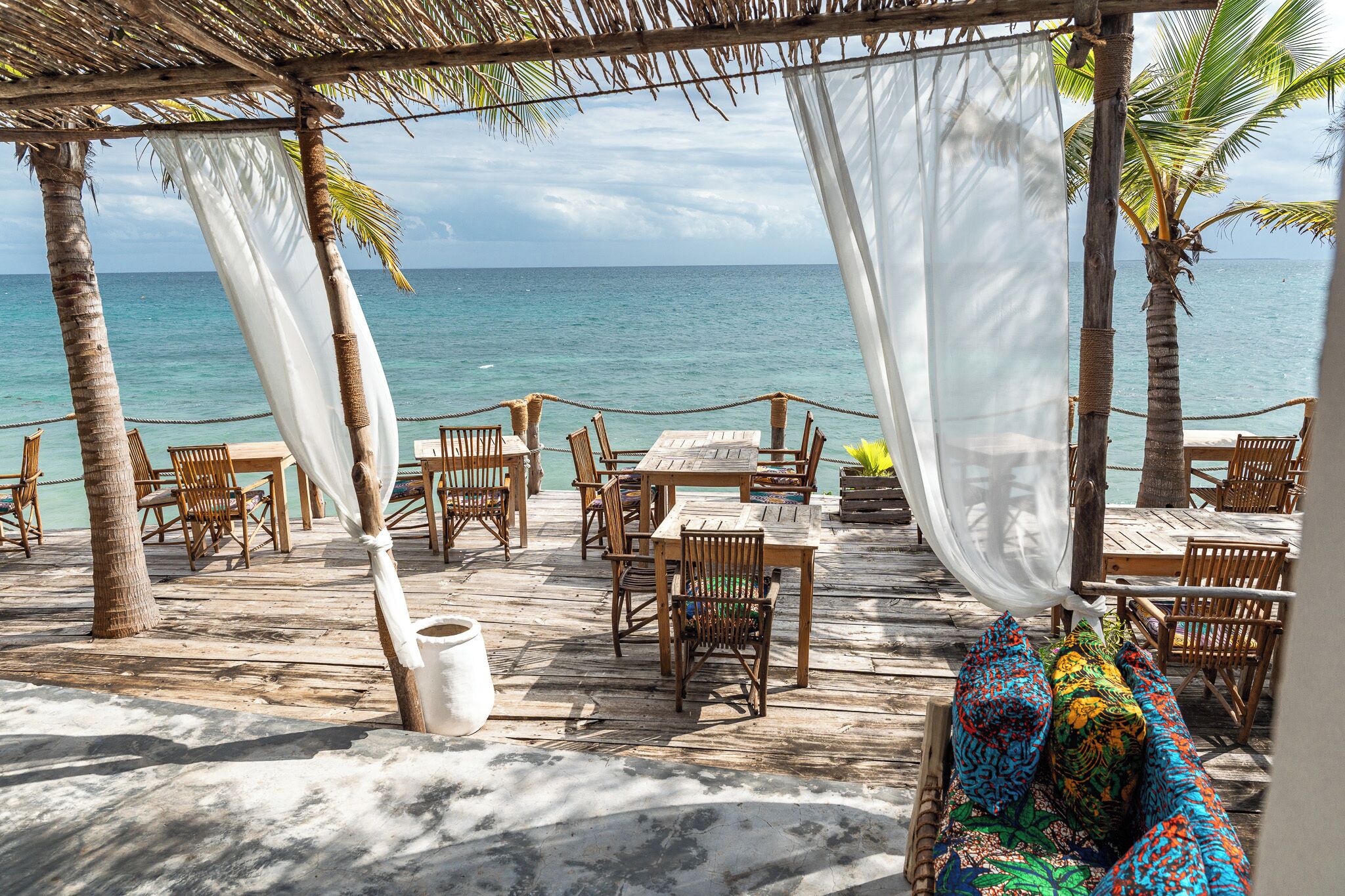 A beachside patio with wooden tables and chairs, a thatched roof, and a view of the ocean.