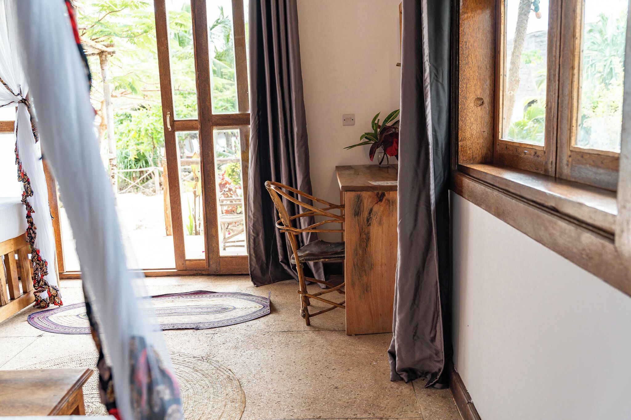 Habitació amb una cadira de fusta, una taula petita amb una planta, una finestra amb cortines i vista a la vegetació exterior.