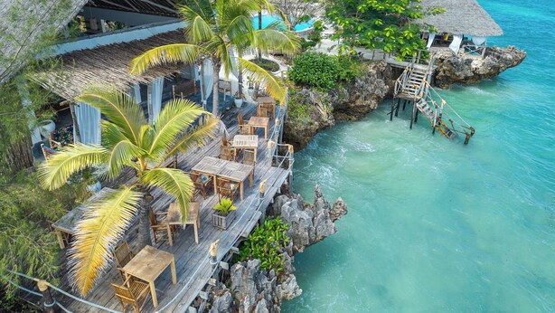 Un resort tropical con edificios de techo de paja, asientos en terraza de madera y un mar turquesa cristalino.