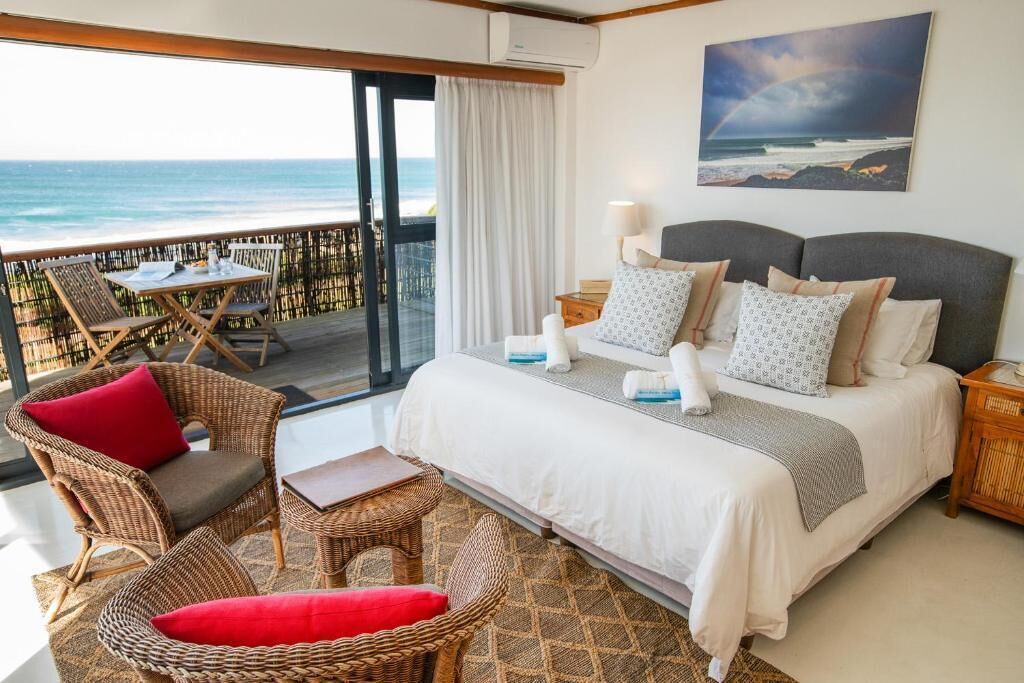 Una camera da letto con un letto, due comodini, un piccolo tavolo, una sedia in vimini e un balcone con vista sull'oceano.