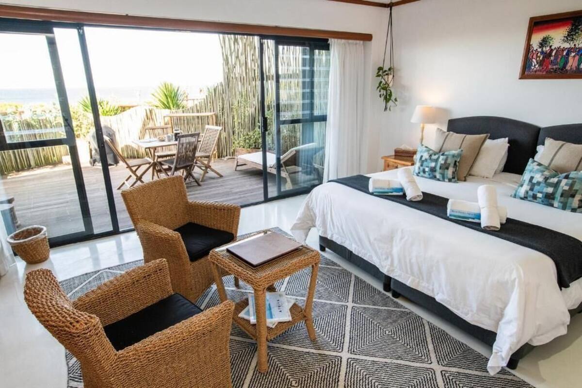 A hotel room with a bed, a small table, wicker chairs, a balcony with a table and chairs, and a view of the sea.