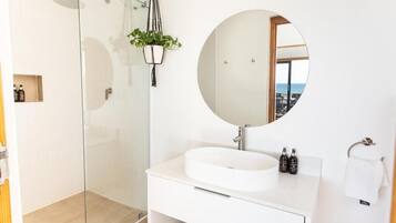 Une salle de bain moderne avec une douche à parois de verre, un miroir rond, un lavabo blanc et une petite étagère contenant des articles de toilette.