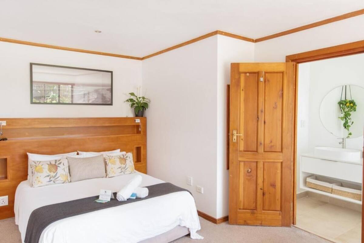 A hotel room with a wooden headboard, a bed with white bedding, a bedside table with a plant, and a bathroom with a white sink and mirror.