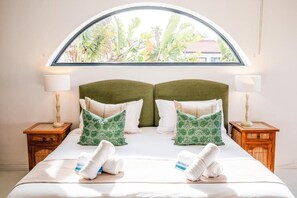 A double bed with two nightstands, a large arched window, and a view of greenery outside.