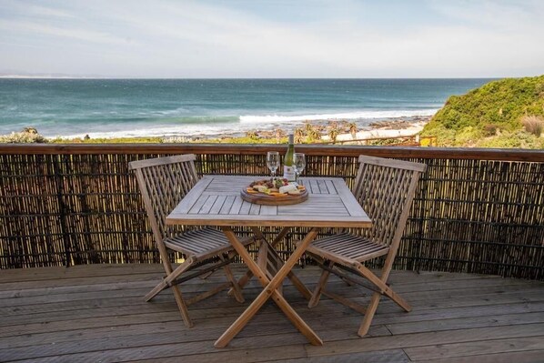 A wooden deck with a table and chairs, overlooking a beach and ocean.
