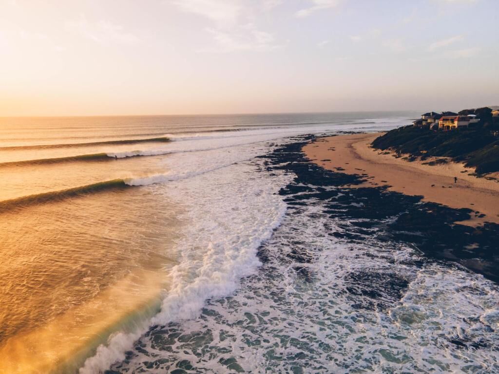 Una playa con olas, una orilla arenosa y edificios al fondo.