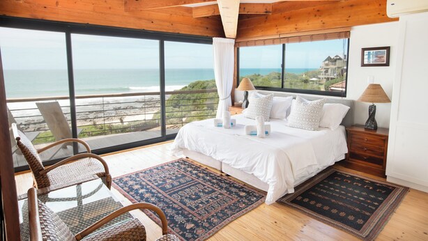 Una camera da letto con un'ampia finestra vista spiaggia, un letto con lenzuola bianche, due abat-jour e una poltrona in vimini.