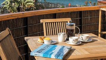 Ein Holztisch mit einem Buch, einer Kaffeekanne und Tassen auf einem Balkon mit Blick auf den Ozean.