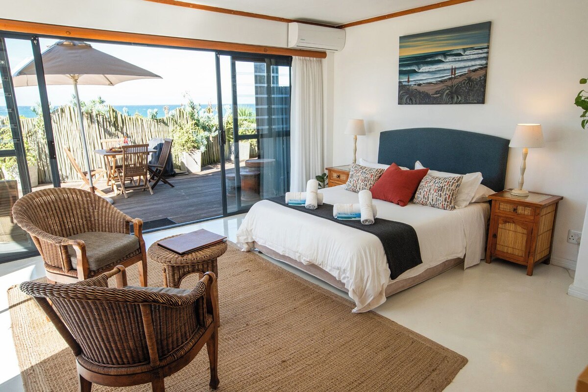 A bedroom with a bed, two nightstands, a chair, a table, and a view of the ocean.