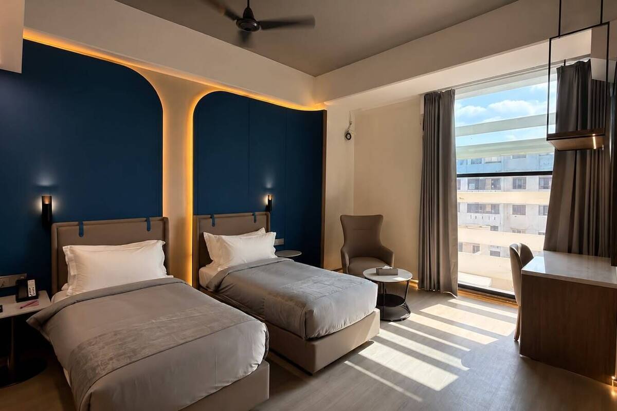 Ein Hotelzimmer mit zwei Betten, einem Schreibtisch, einem Sessel, einem kleinen Tisch und einem großen Fenster mit Vorhängen.