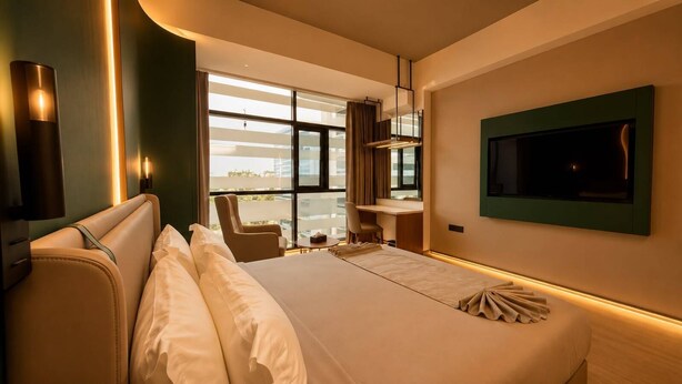 A hotel room with a bed, a desk, a chair, a TV, and a view of the city. - HOTEL NATANYA (Thanjavur)