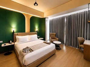A hotel room with a bed, bedside tables, a desk, a chair, and a large window with curtains. - HOTEL NATANYA (Thanjavur)