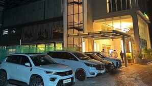 A building with a modern design, featuring a prominent vertical structure and a glass facade. There are several white vehicles parked in front. - HOTEL NATANYA (Thanjavur)