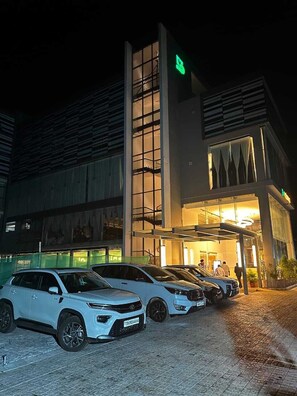 A building with a modern design, featuring a prominent vertical structure and a glass facade. There are several white vehicles parked in front. - HOTEL NATANYA (Thanjavur)