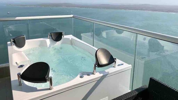 Un balcon fermé par des baies vitrées, doté d’un jacuzzi et de deux fauteuils, offrant une vue sur une étendue d’eau et sur la terre ferme au loin.